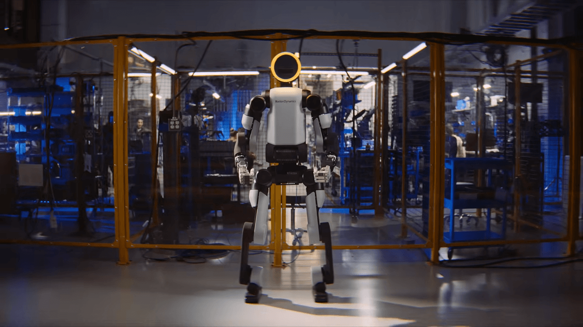 A humanoid robot stands in a workshop surrounded by yellow safety barriers.