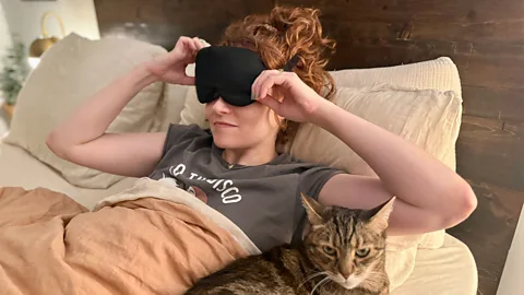 Woman pulling off sleep mask while waking naturally in bed with cat beside her